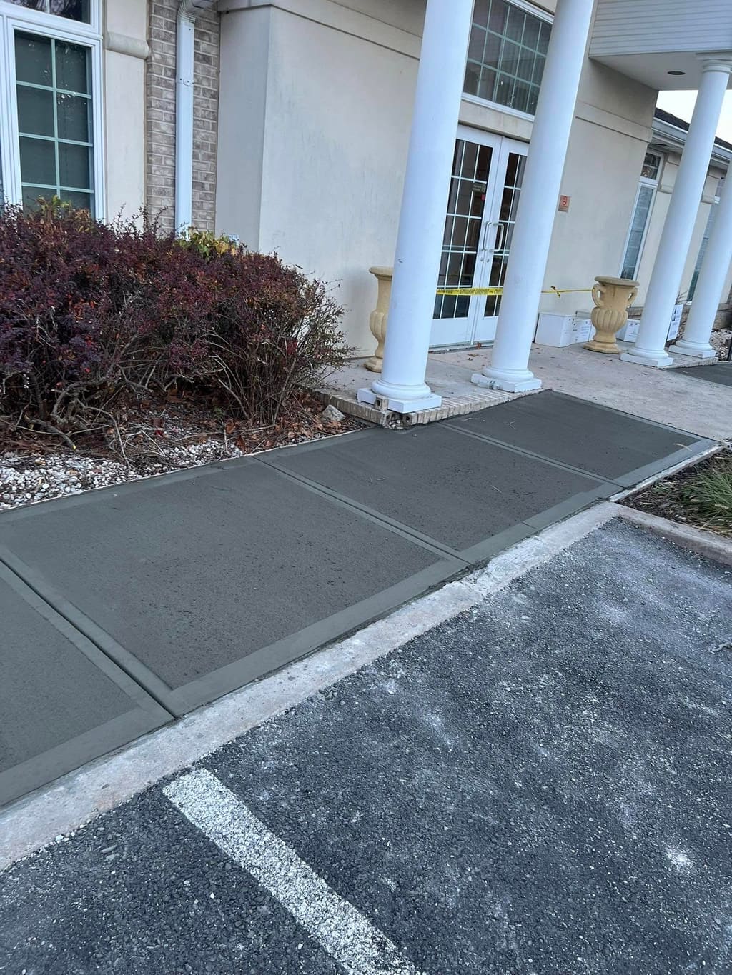 Finished concrete walkway leading to a columned building entrance beside an asphalt parking lot.