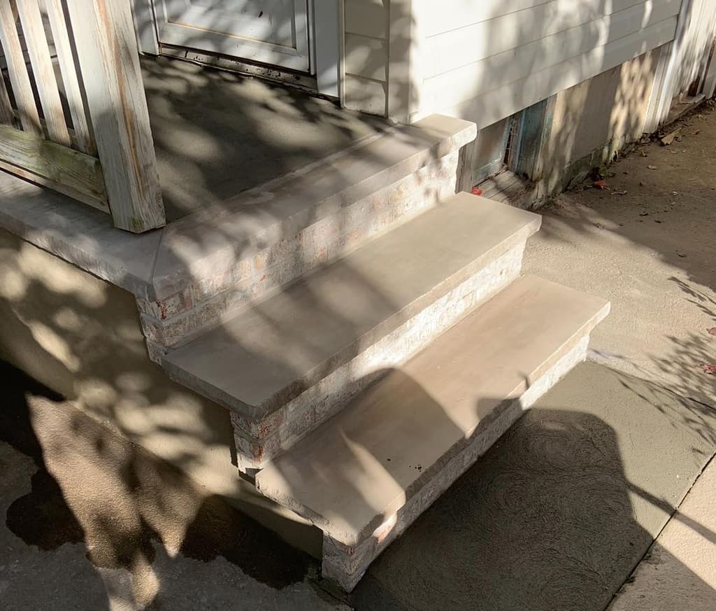 Concrete steps with stone-veneer risers at a side entrance, partly shaded by tree shadows.