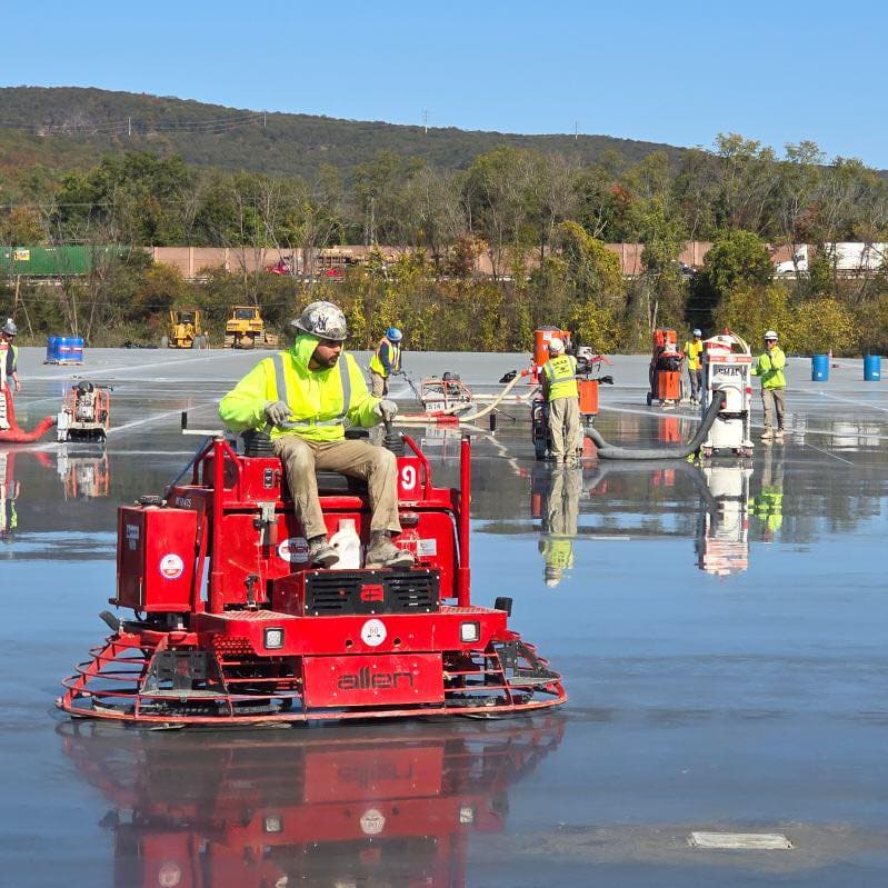 Worker on a ride-on power trowel finishing a large freshly poured concrete slab outdoors.
