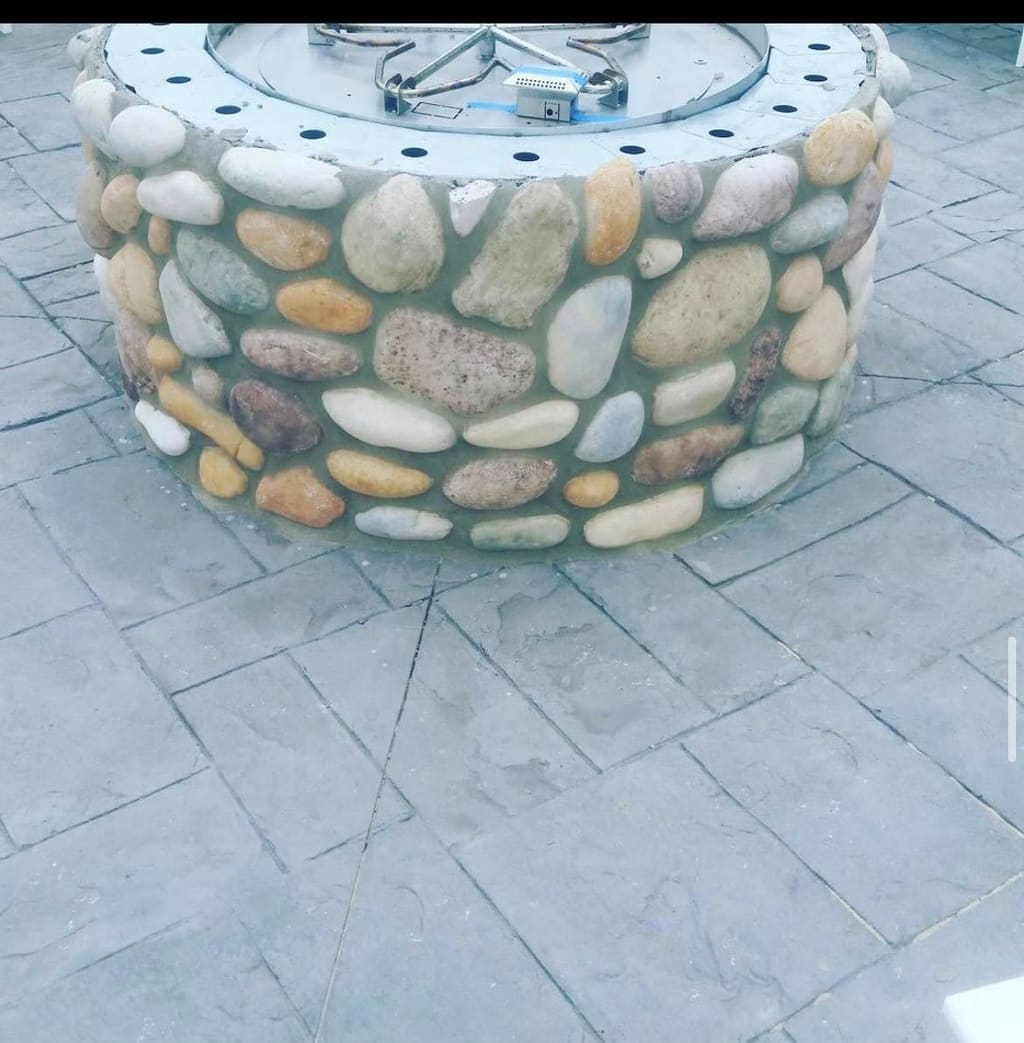 Round fire pit faced with multicolored river-rock veneer set on a stamped concrete patio.