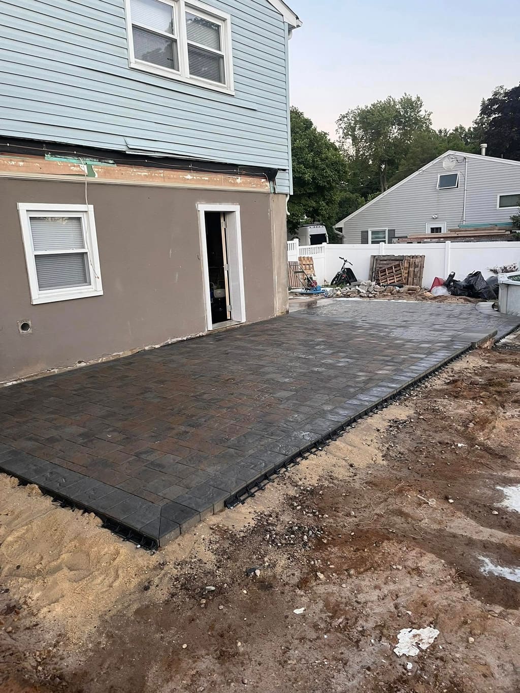 Newly installed gray paver patio with a step against a house, surrounded by unfinished soil.