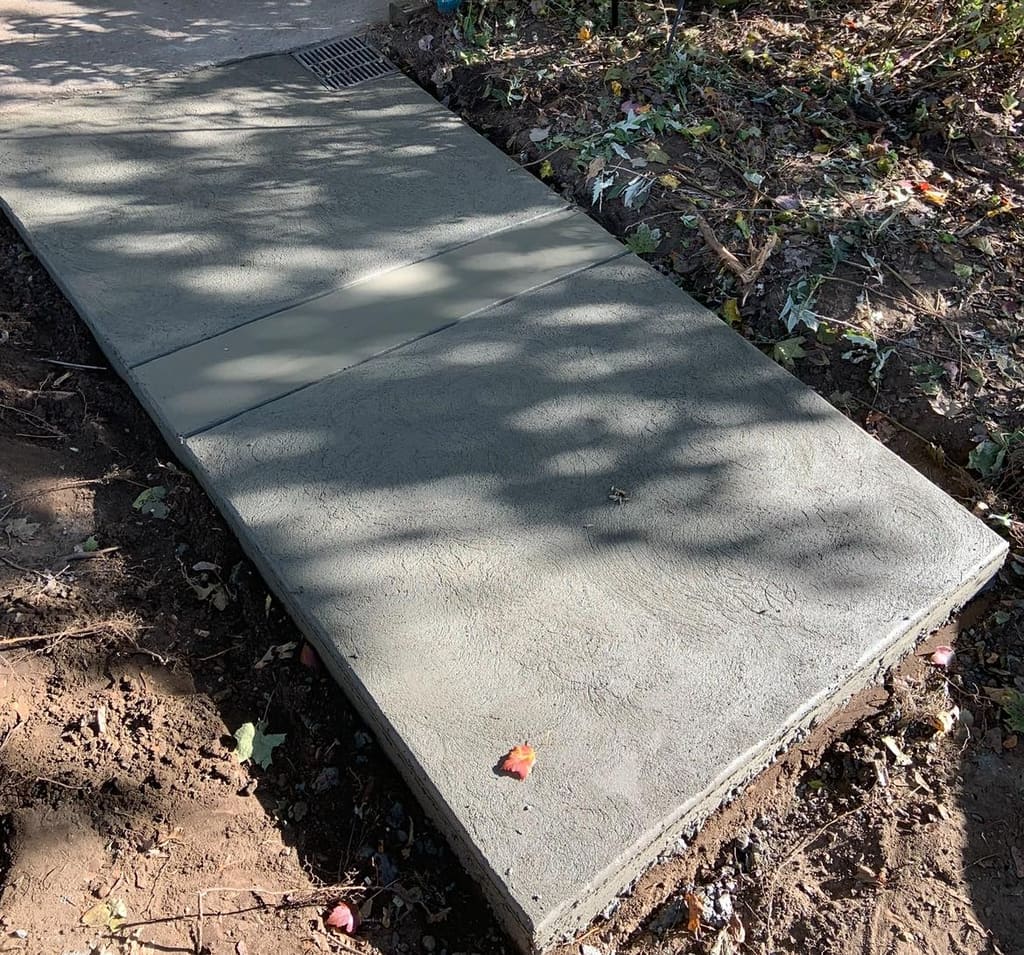 Newly poured smooth concrete walkway with a drain grate set between dirt garden beds.