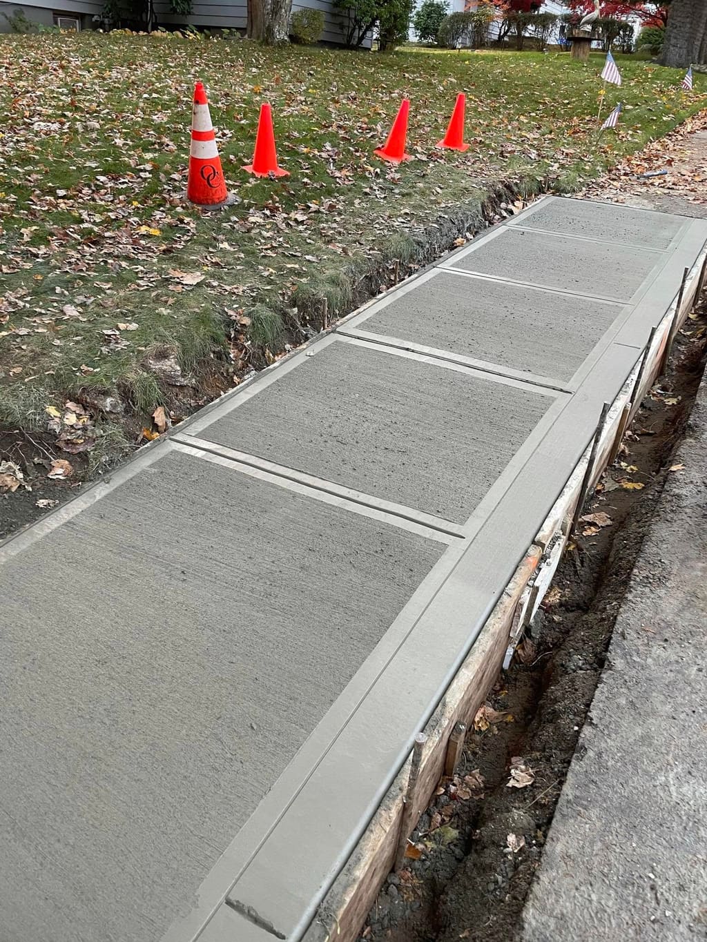 Freshly poured sectioned concrete sidewalk with wood forms still in place and safety cones nearby.