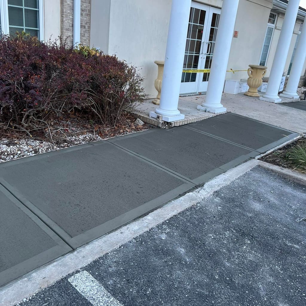 Finished concrete walkway leading up to a columned building entrance beside a paved parking area.