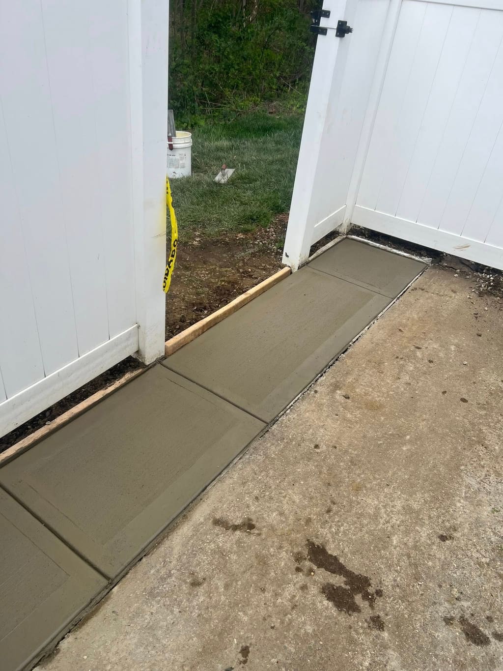 Freshly poured concrete walkway pad set between a white vinyl fence gate and an existing path.