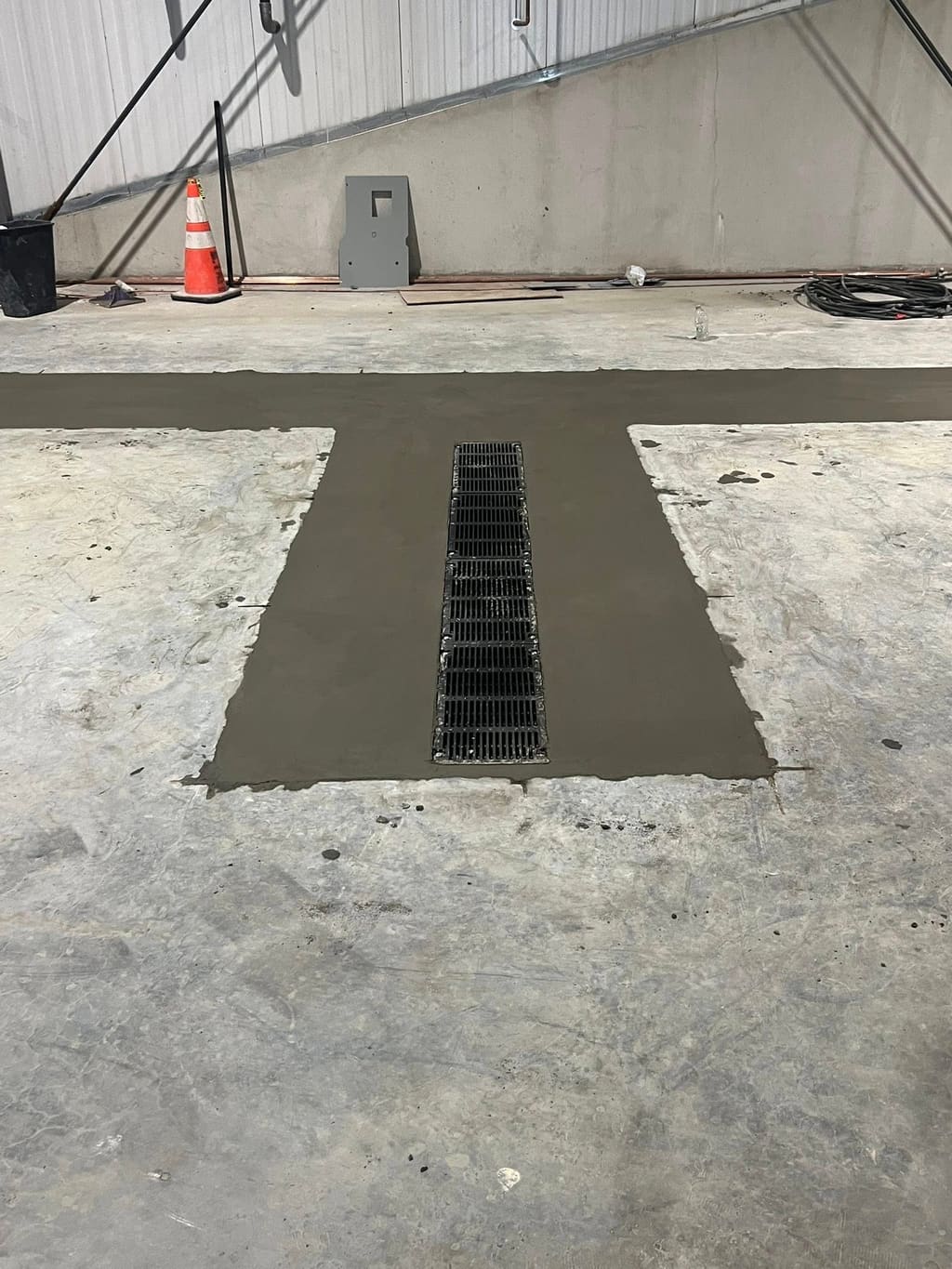 Freshly poured concrete slab patch surrounding a metal trench drain grate on a warehouse floor.