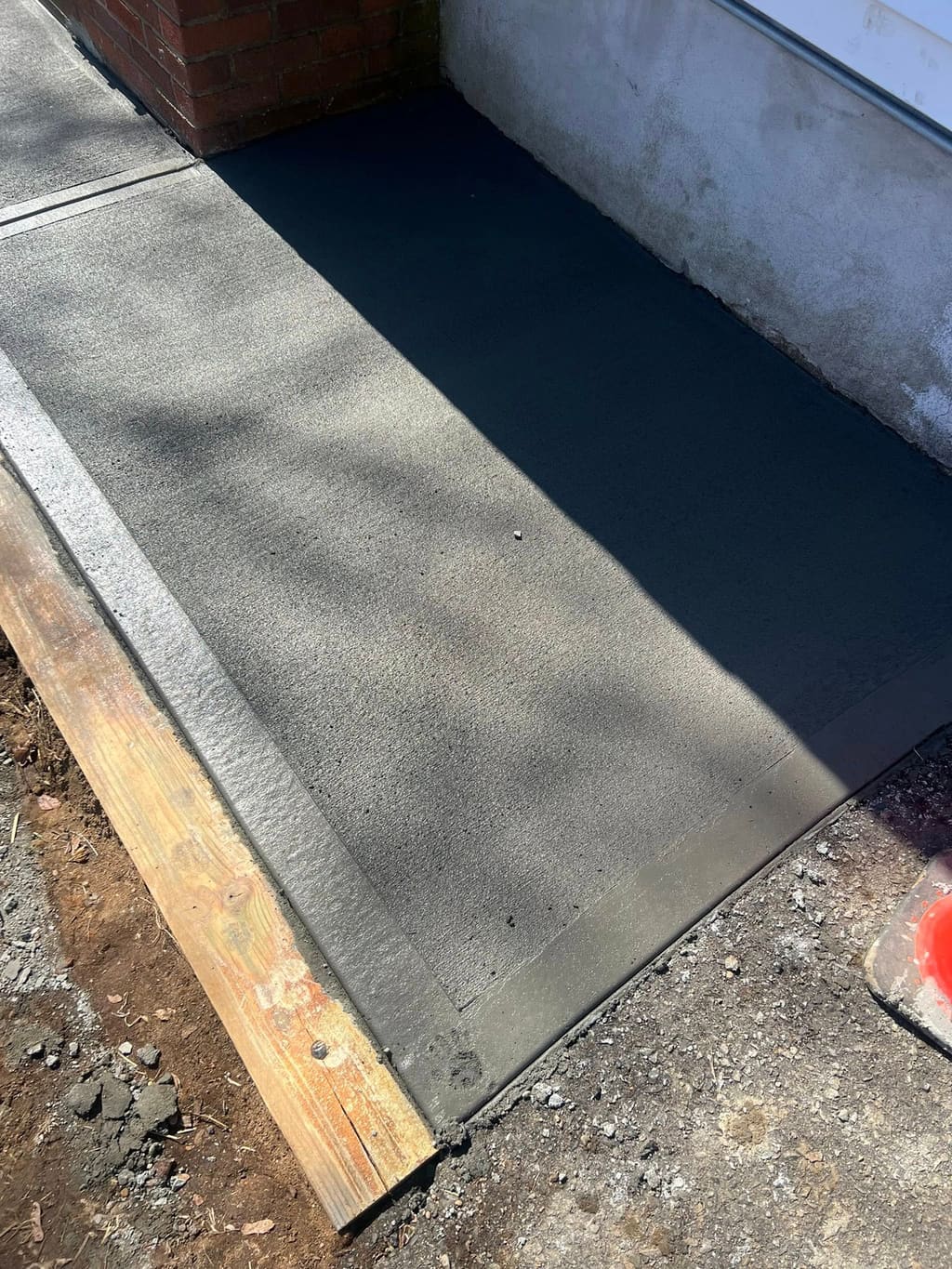 Newly finished gray concrete walkway running alongside a brick building wall with a wood form board.