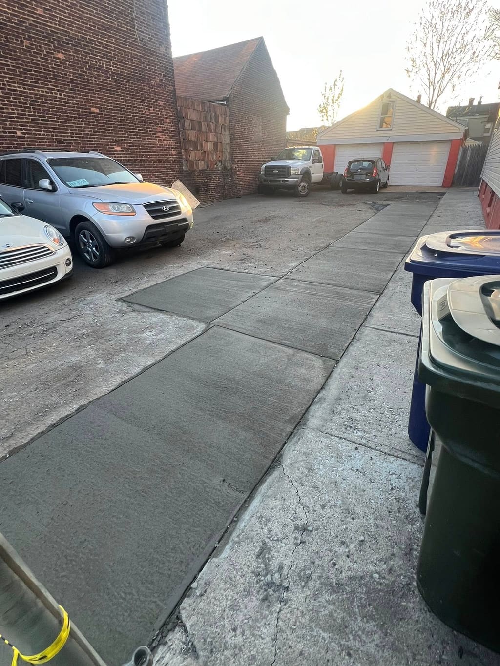 Newly poured concrete slab sections in a backyard parking driveway with cars and garages behind.