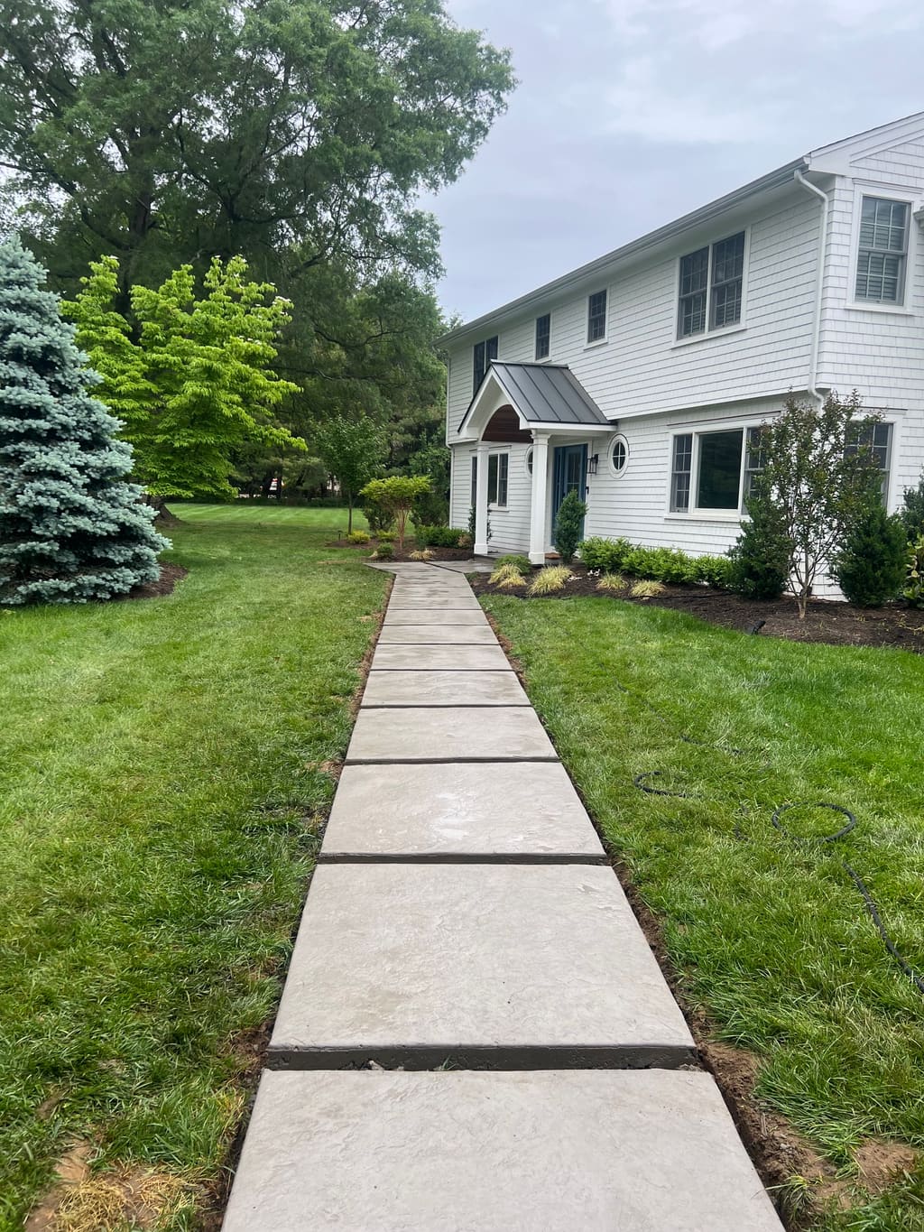 Long straight concrete walkway with control joints running across a lawn beside a white house.