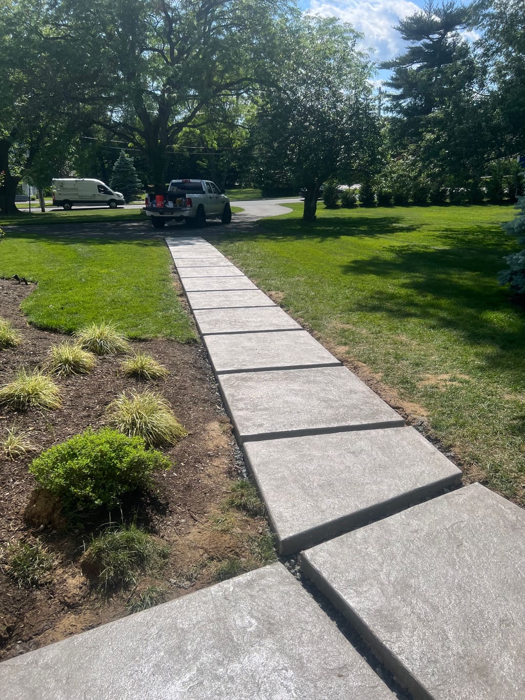Long sectioned concrete walkway with control joints crossing a green lawn toward parked equipment.