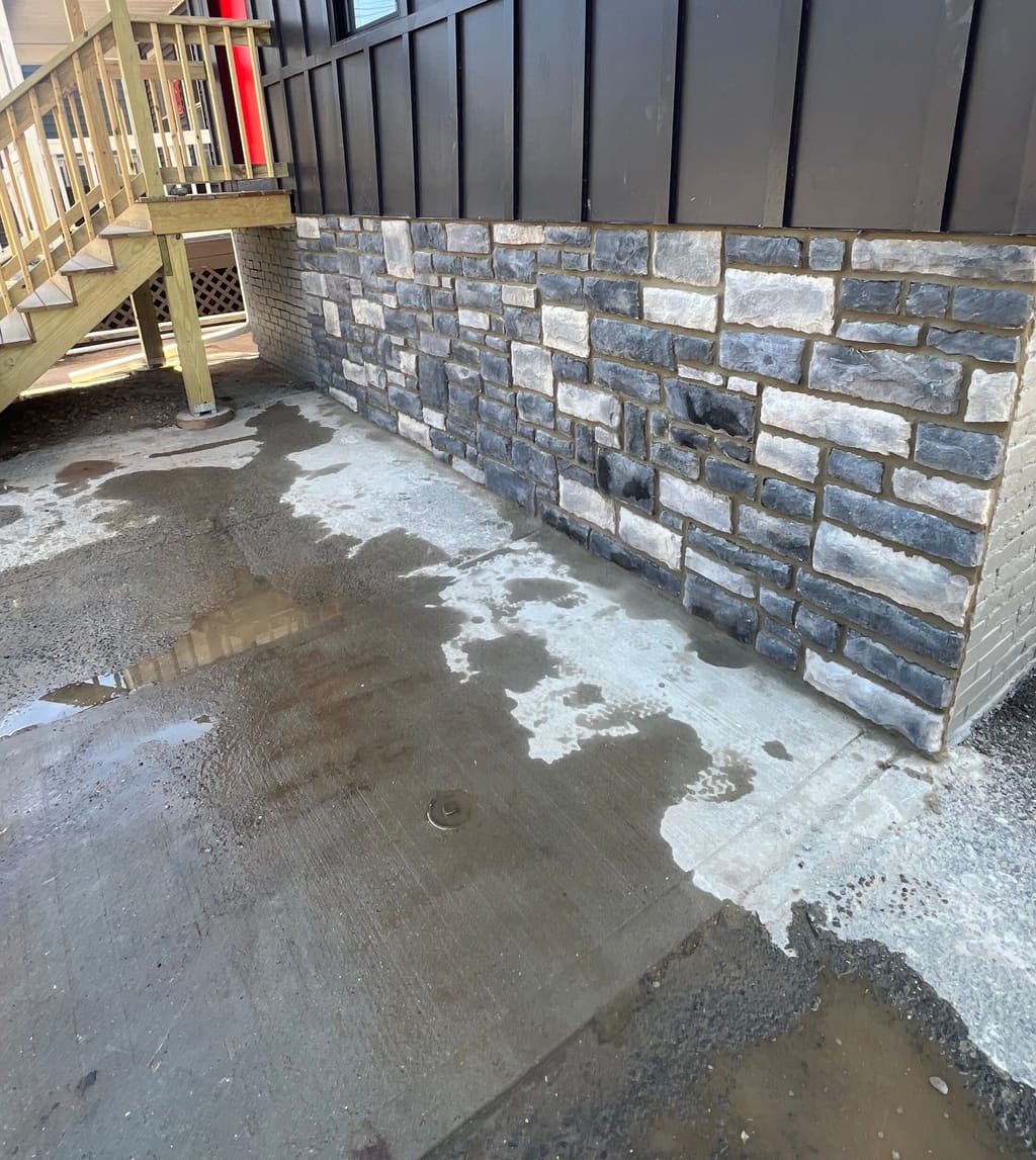 Gray ledgestone veneer applied to the foundation wall of a dark board-and-batten building beside a wood stair.