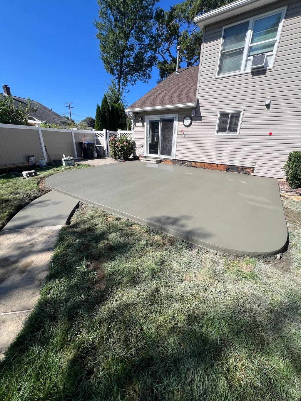 Large freshly poured concrete patio with a rounded edge behind a house, set into the lawn by a vinyl fence.