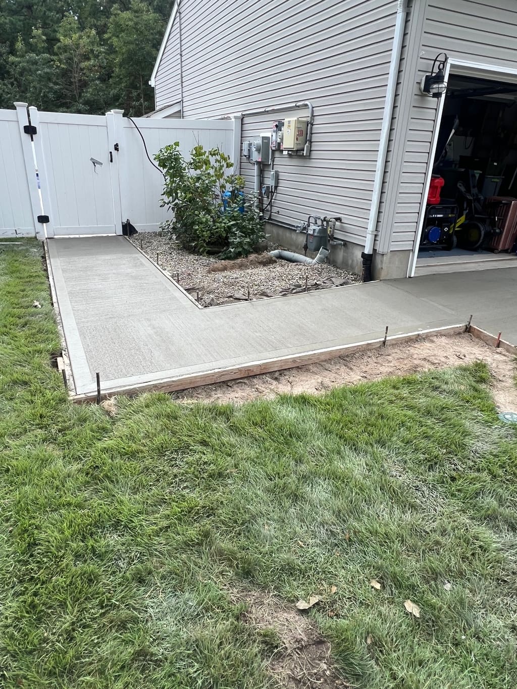 New concrete side-yard pad poured beside a garage and utility meters, framed by a vinyl fence and lawn.