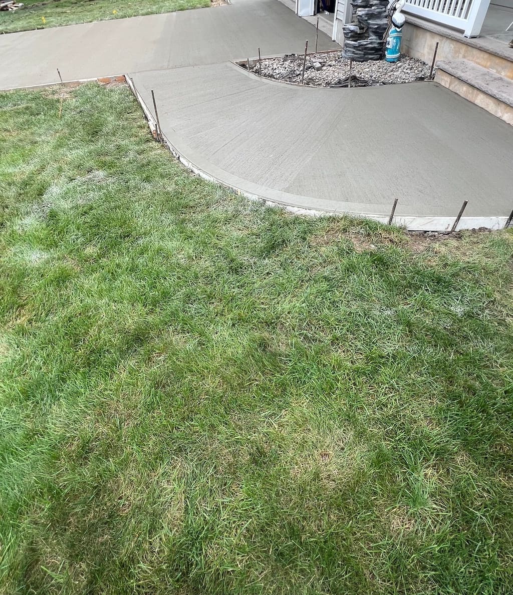 Freshly poured curved concrete entry pad and walkway in front of a porch, with stakes still in place.
